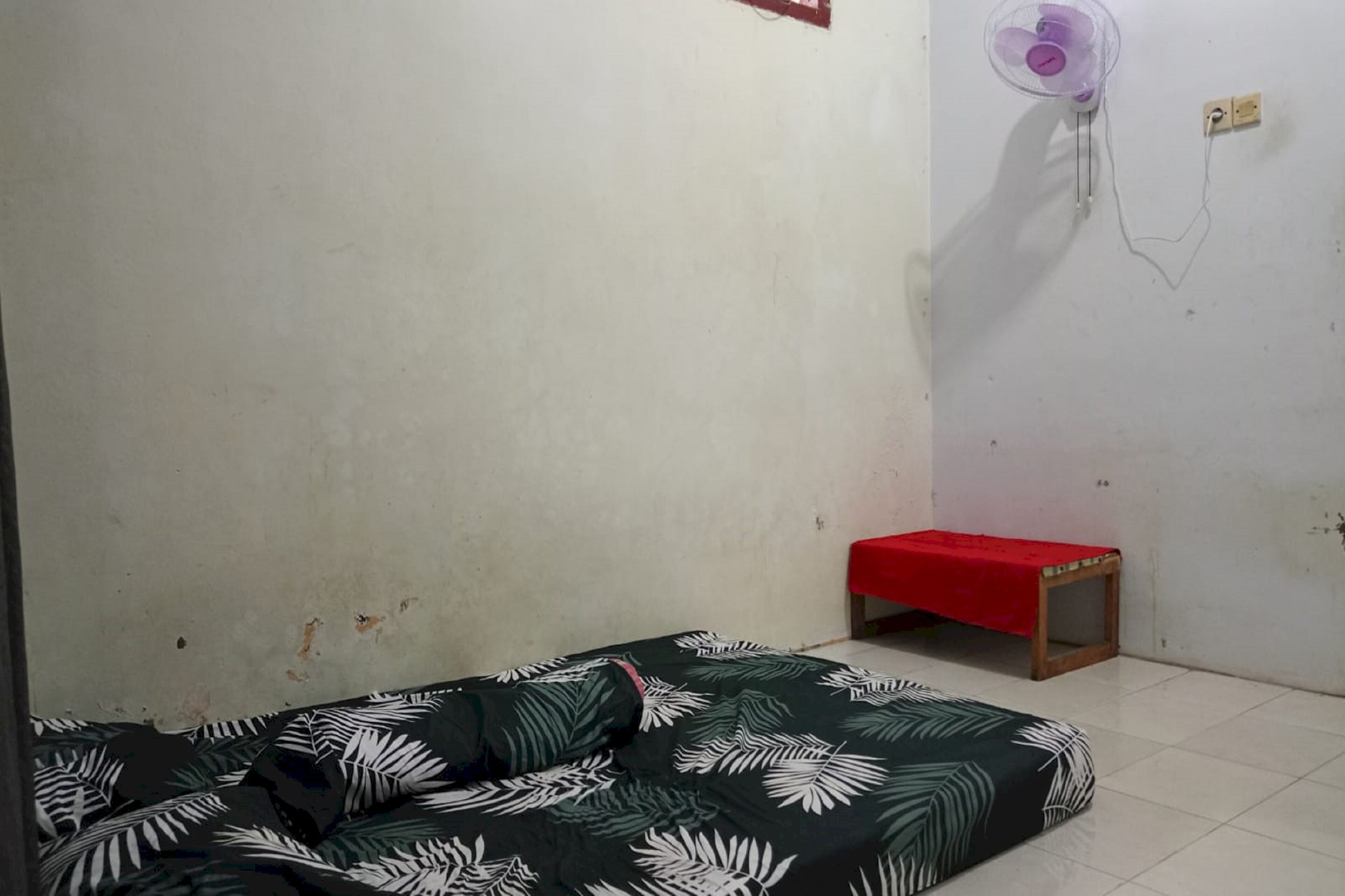 Una habitació amb un llit, una taula petita amb tela vermella i un ventilador fixat a la paret.