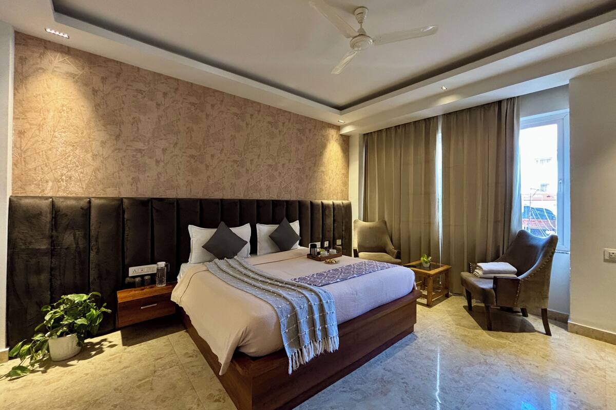 Een moderne hotelkamer met een groot bed, een plafondventilator, een zithoek met een stoel en een klein tafeltje, en uitzicht buiten door een raam.