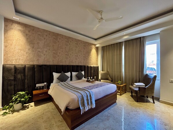 A modern hotel room with a large bed, a ceiling fan, a seating area with a chair and a small table, and a view of the outside through a window.