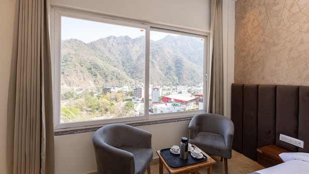 A hotel room with two armchairs, a small table with a tray, a bed, and a large window offering a mountain view. - Vyom Boutique Hotel (Narendranagar)