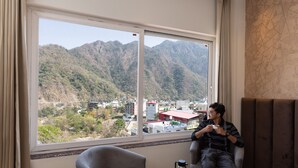 A room with a large window offering a view of mountains and a town. There is a chair, a table with a teapot, and a cup. - Vyom Boutique Hotel (Narendranagar)