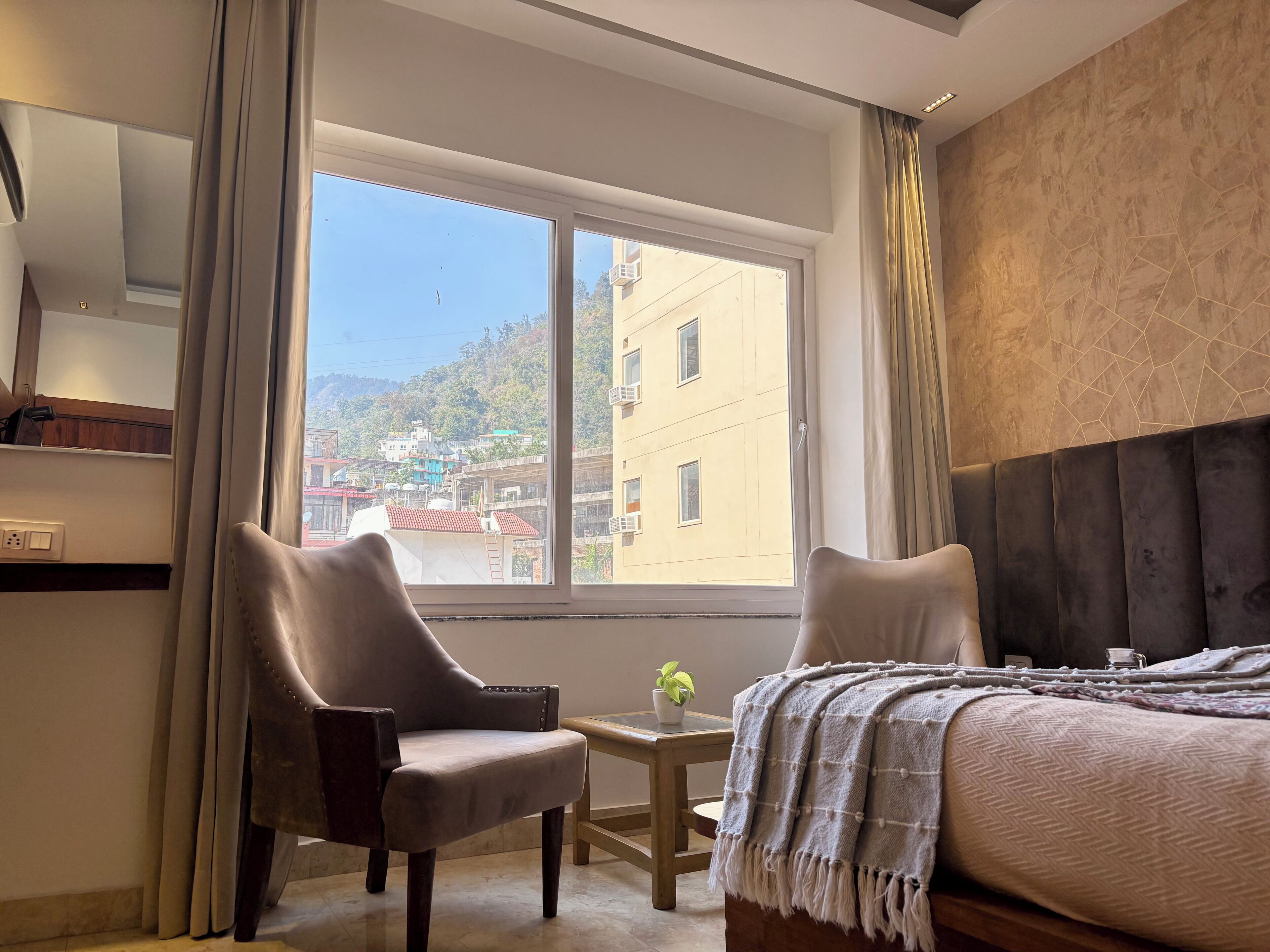 A hotel room with a large window, a bed, a chair, a small table, and a potted plant.