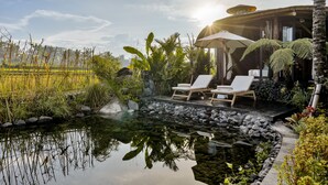 Tranquila zona de piscina al aire libre con sillas de descanso y un área de estar cubierta, rodeada de exuberante vegetación y un cielo despejado.