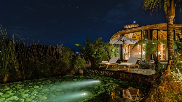 Vista nocturna de un resort con piscina, sillones de descanso y un espacio exterior bien iluminado.