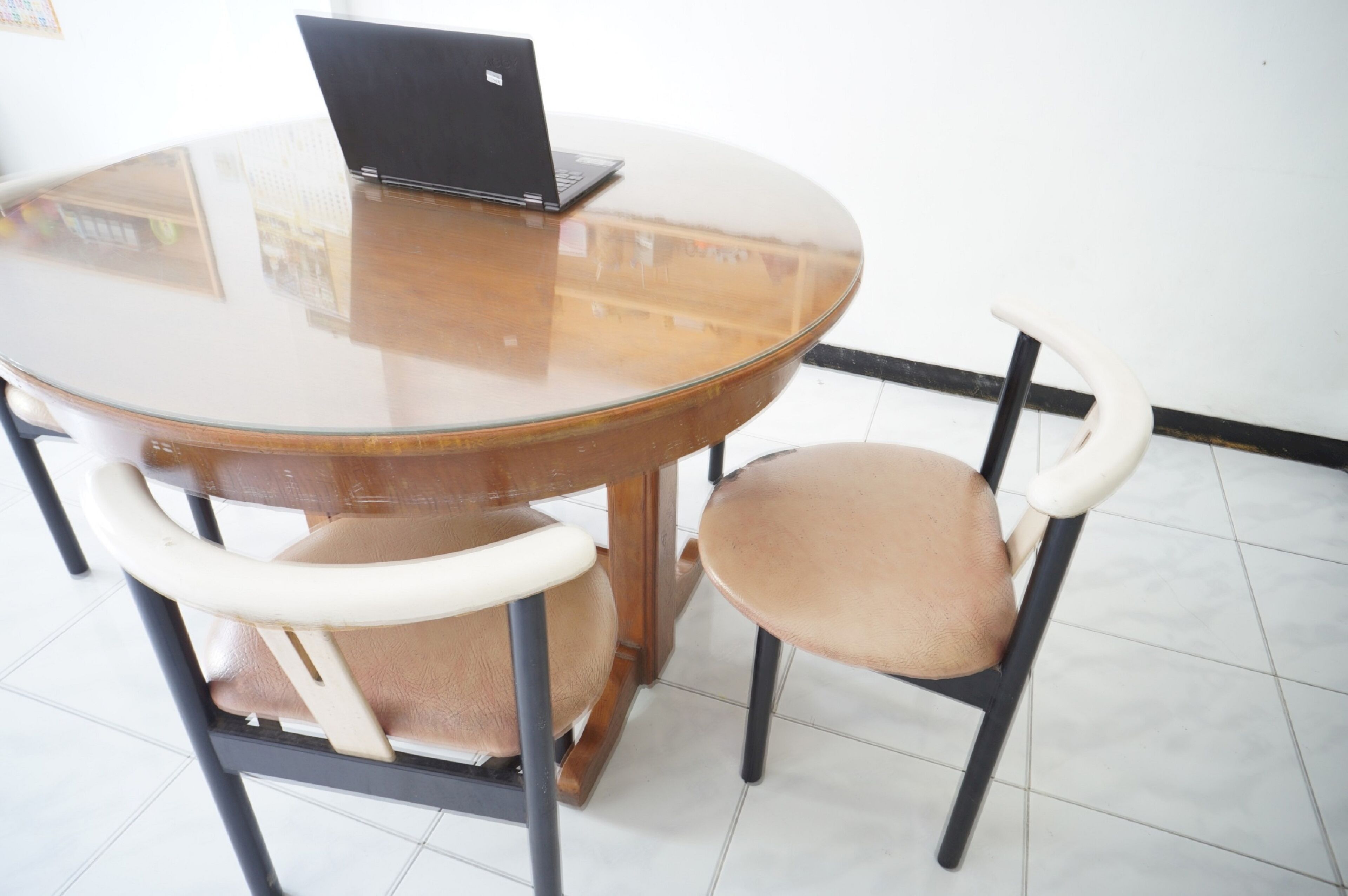 Una mesa redonda de vidrio con un ordenador portátil encima, rodeada por dos sillas con asientos acolchados y estructura metálica.