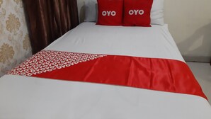 Een bed met een wit-rode spreideken, twee rode kussens met de tekst 'OYO' erop en een bruin gordijn.