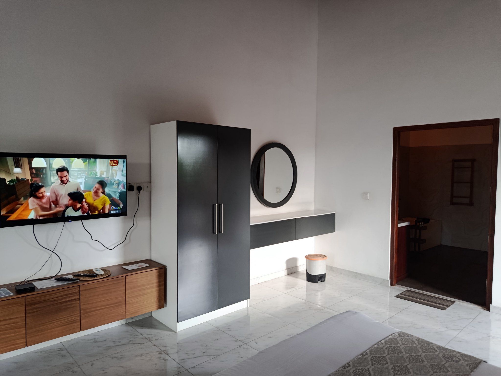 A modern living room with a flat-screen TV mounted on the wall, a dark-colored cabinet with a mirror above it, and a small trash bin.