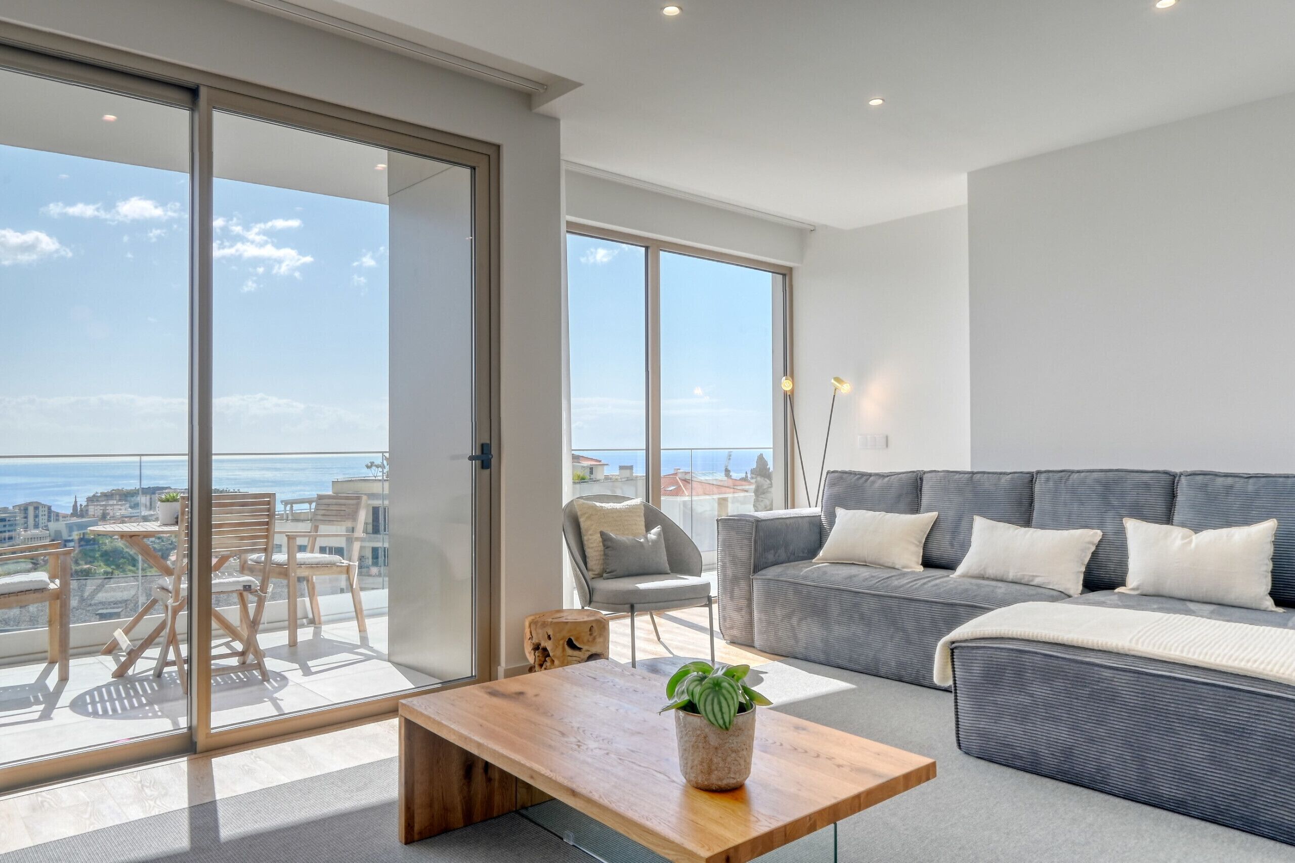 A modern living room with a grey sofa, a wooden coffee table, and a balcony with a view of the city and sea.