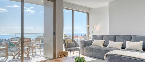 A modern living room with a grey sofa, a wooden coffee table, and a balcony with a view of the city and sea.