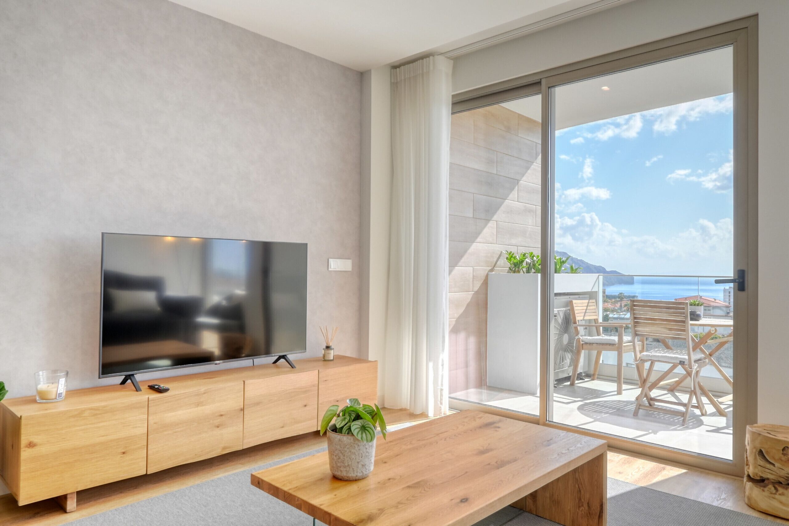 Un salon moderne avec un téléviseur à écran plat posé sur une console en bois, une table basse et une vue sur un balcon aménagé avec du mobilier d’extérieur.
