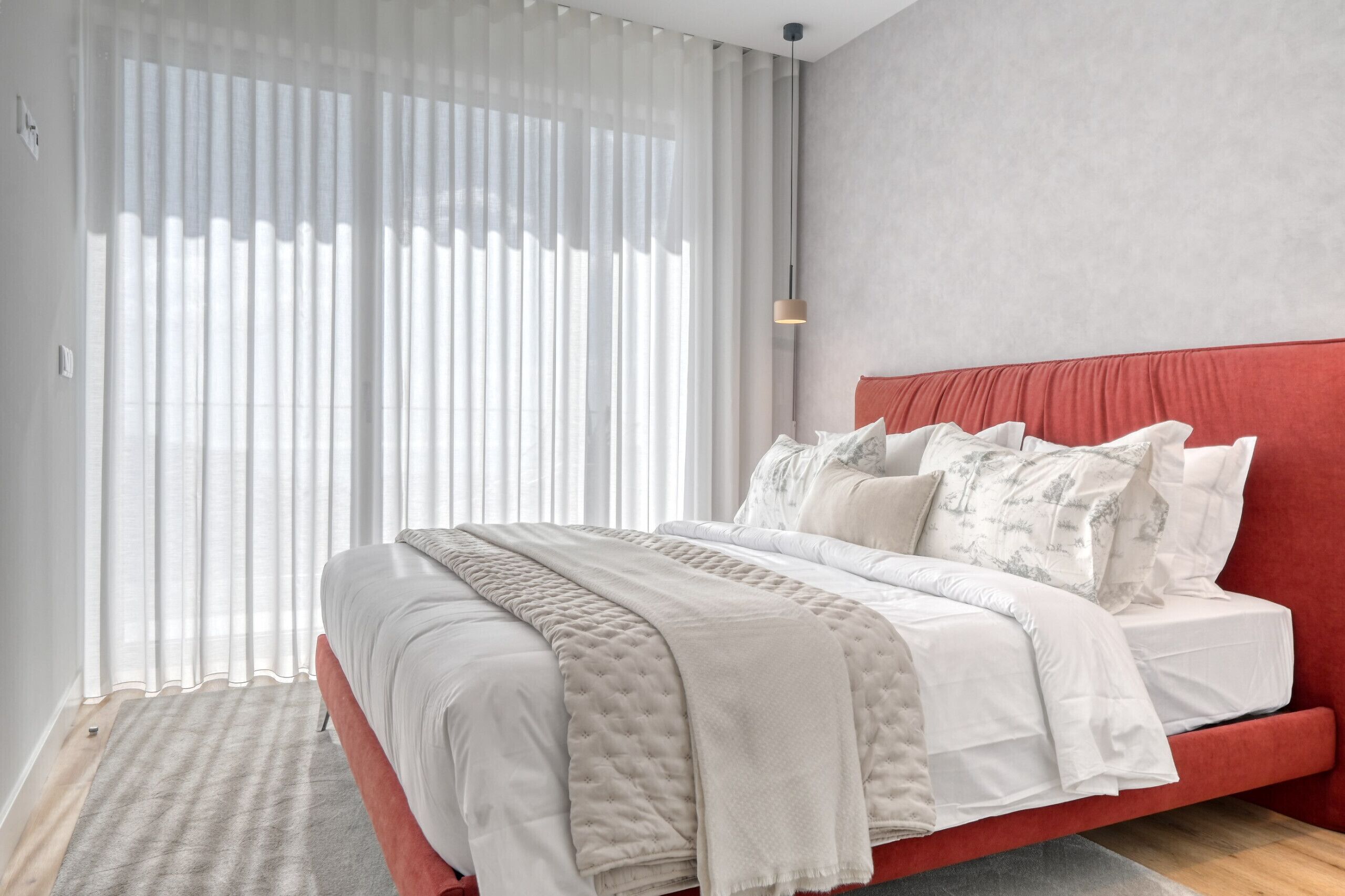Ein modernes Schlafzimmer mit einem roten Kopfteil, einem großen Bett mit weißer Bettwäsche und durchscheinenden Vorhängen, die natürliches Licht hereinlassen.