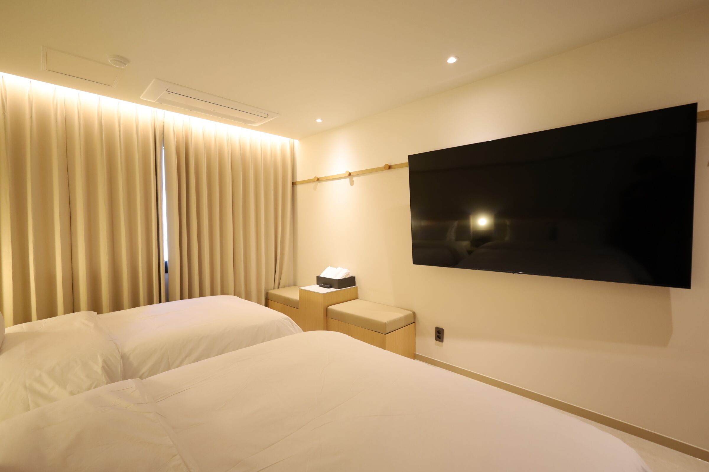 A hotel room with two beds, a flat-screen TV mounted on the wall, a bedside table with a lamp, and a window with curtains.