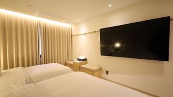 A hotel room with two beds, a flat-screen TV mounted on the wall, a bedside table with a lamp, and a window with curtains.