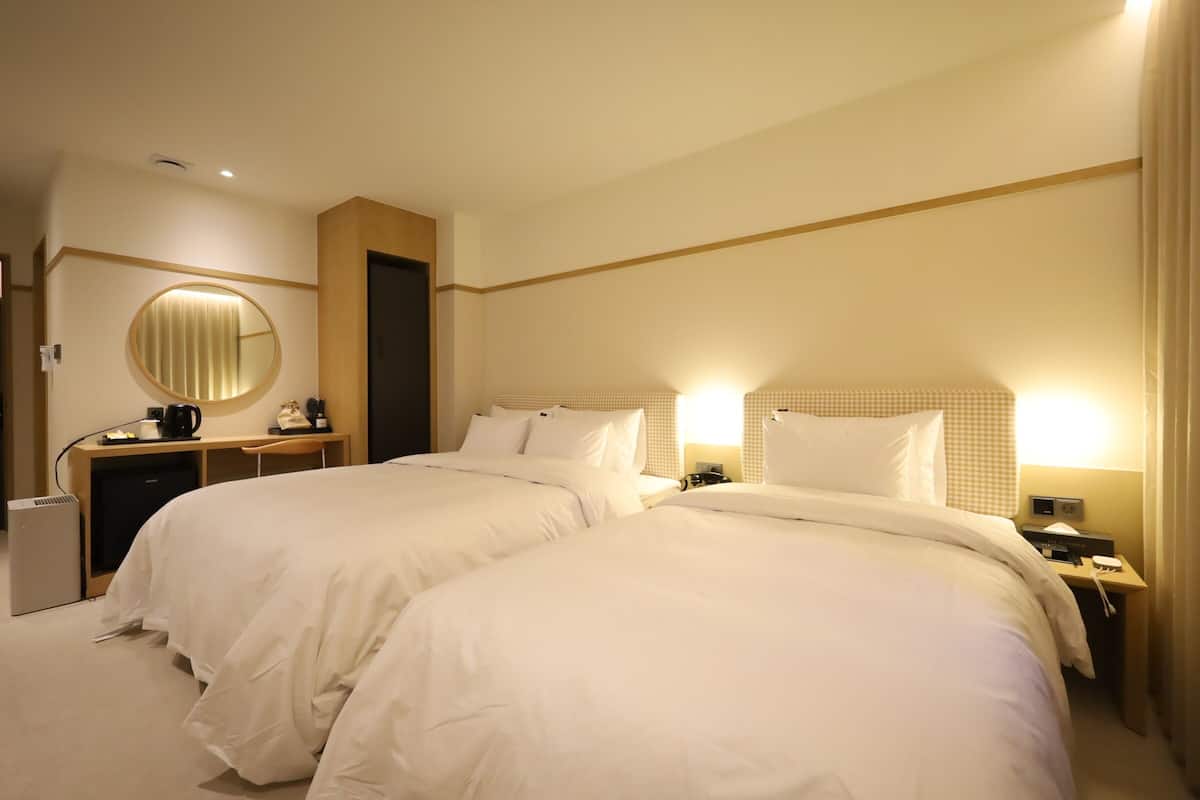 A hotel room with two beds, a desk with a lamp, a mirror, and a small refrigerator.