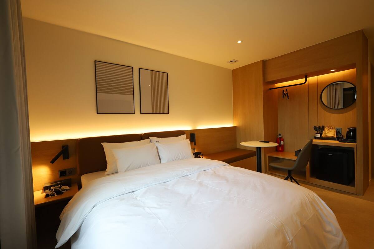 A hotel room with a large bed, bedside tables, a desk, a chair, a small refrigerator, a round mirror, and a wall-mounted shelf.