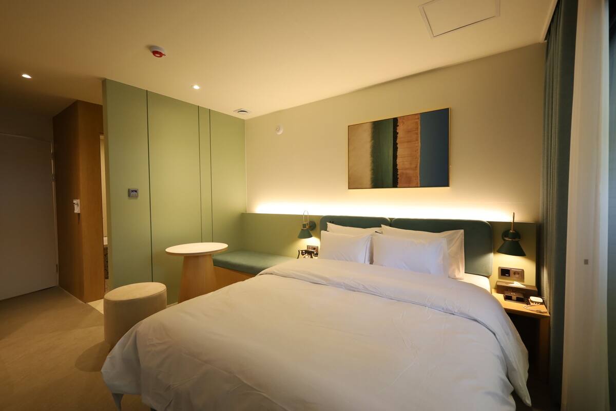 A hotel room with a large bed, a bedside table, a nightstand, a green wall, and a painting.