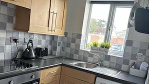 A compact kitchen with wooden cabinets, a built-in oven, a washing machine, and a window with a view of greenery outside. - Black Door Malt - Huku Kwetu (Dunstable)