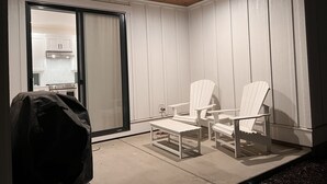 Une terrasse couverte avec deux fauteuils Adirondack blancs, un barbecue et une porte-fenêtre coulissante donnant sur la cuisine.