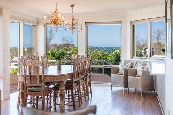 A dining area with a round wooden table and chairs, a chandelier, and large windows offering a view of the outdoors. - Sandrift One - Oceanstays (Yamba)