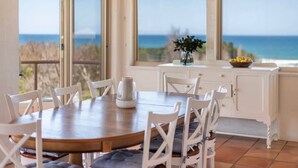 Un coin repas avec une table en bois et des chaises blanches, de grandes fenêtres donnant sur l’océan, et un meuble blanc sur lequel est posé un saladier rempli de citrons.