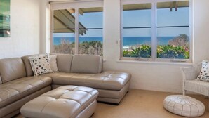 Un salon moderne avec un canapé d’angle beige, un pouf rond et un fauteuil en osier. Deux grandes fenêtres offrent une vue sur l’océan et la végétation.