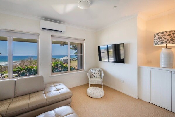 A room with a beige leather sofa, a white chair, a large window with a view of the sea, a mounted TV, and a white radiator. - Sandrift Two - Oceanstays (Yamba)
