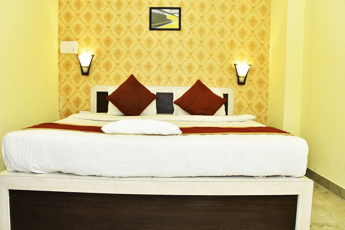 A neatly made bed with white bedding and two brown pillows, a wall-mounted light fixture, and a framed picture above the bed.