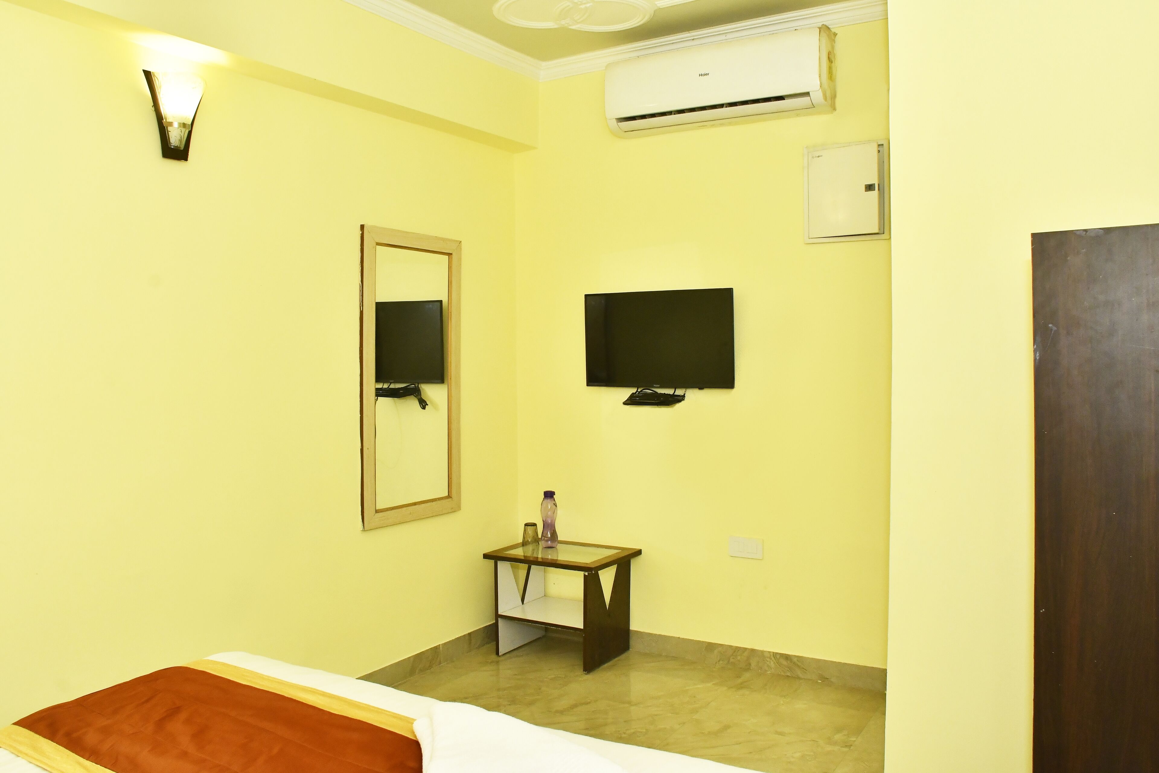 A hotel room with a bed, a bedside table with a lamp, a TV mounted on the wall, and an air conditioning unit.
