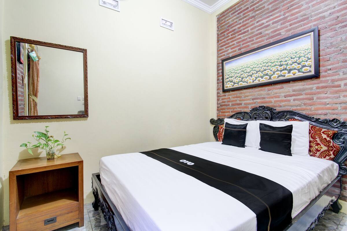 A bedroom with a brick wall, a bed with a black and white bedspread, a wooden nightstand with a plant, and a mirror.