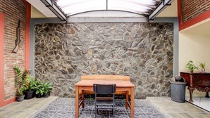 Un mur de pedra, una taula de fusta amb cadires i plantes en testos a un pati.