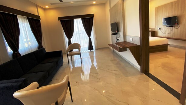A modern hotel room with a flat-screen TV mounted on the wall, a ceiling fan, a sofa, a chair, and a wooden bench. - The Grand Sarovar Inn and Suites (Rajgurunagar)