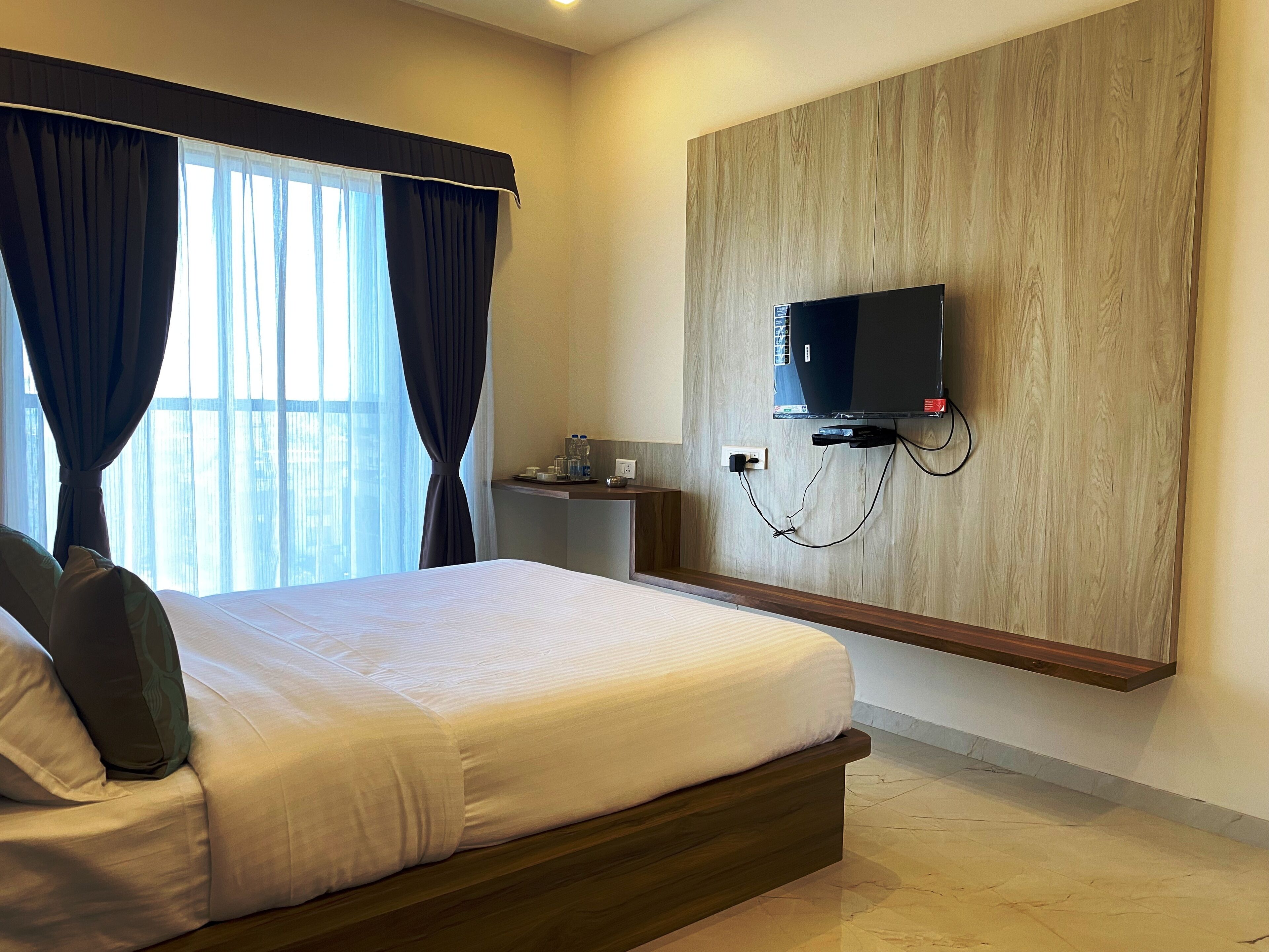 A hotel room with a large bed, a TV mounted on the wall, a small table with a water bottle, and a window with curtains.