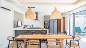 A modern kitchen with a central island, two hanging wicker chandeliers, and a dining area with a wooden table and wicker chairs. - Modern 3 Bedroom Villa - KBR16 (Koh Samui)