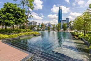 A serene outdoor pool area with a wooden deck, surrounded by lush greenery and modern buildings in the background.
