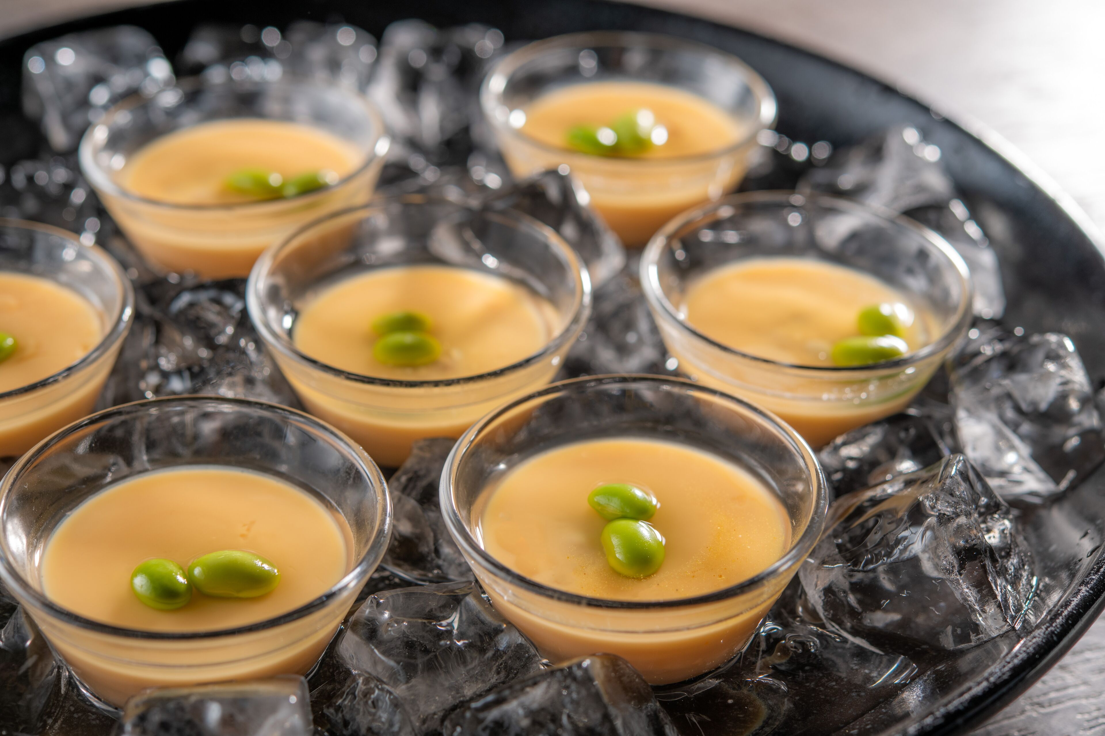 Una bandeja con vasitos de vidrio que contienen un postre cremoso, cada uno decorado con un guisante verde, sobre una cama de cubitos de hielo.