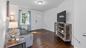 A living room with a sofa, a lamp, a TV on a brick wall, and a door leading to another room. - $ Cozy 1BR 1BA Apartment: Near Fort Gregg-Adams $ (Hopewell)