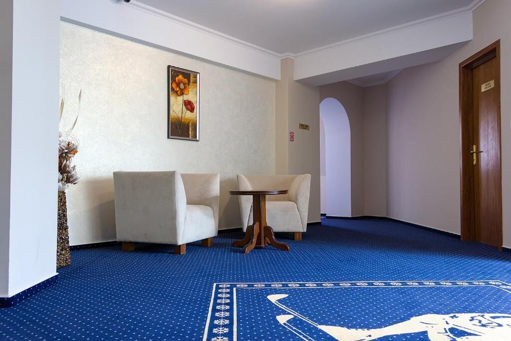 A hotel lobby with a blue carpet, two white armchairs, a small round table, and a framed artwork on the wall.