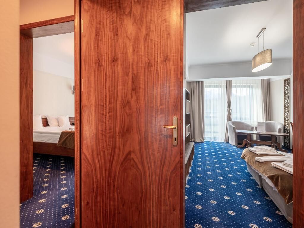 A hotel room with a wooden door, a bed with a blue patterned bedspread, a desk with a chair, and a hanging light fixture.