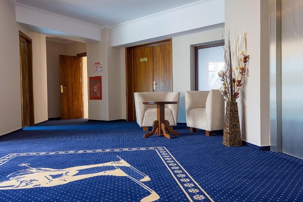 Um saguão de hotel com carpete azul, duas poltronas brancas, uma mesa redonda pequena e um vaso decorativo.