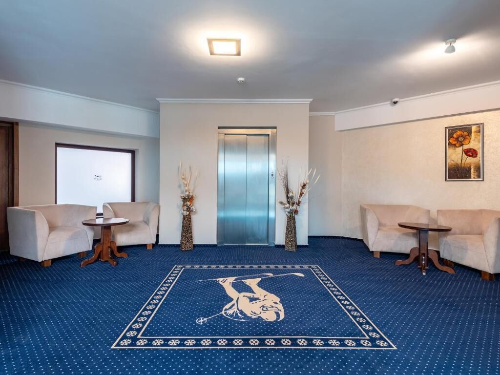 A hotel lobby with a blue carpet, two sofas, a small table, an elevator, and a painting on the wall.