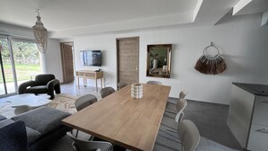 A modern living room with a wooden dining table, grey upholstered sofa, and a television on a stand.