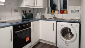 Een kleine keuken met witte kastjes, een inbouwoven, een wasmachine en een raam met potplanten.