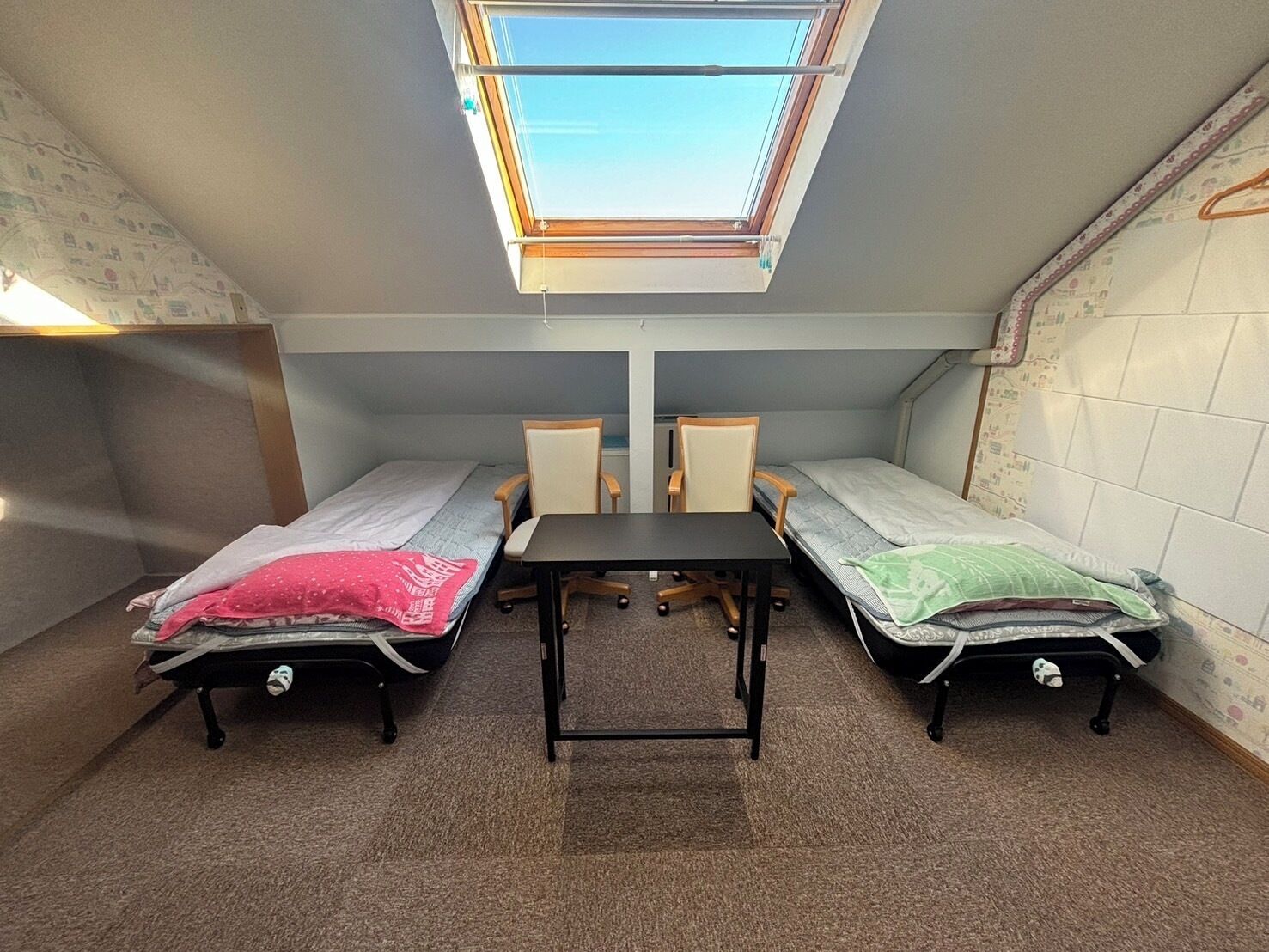 Habitación tipo dormitorio con dos camas individuales, una mesita y un tragaluz.
