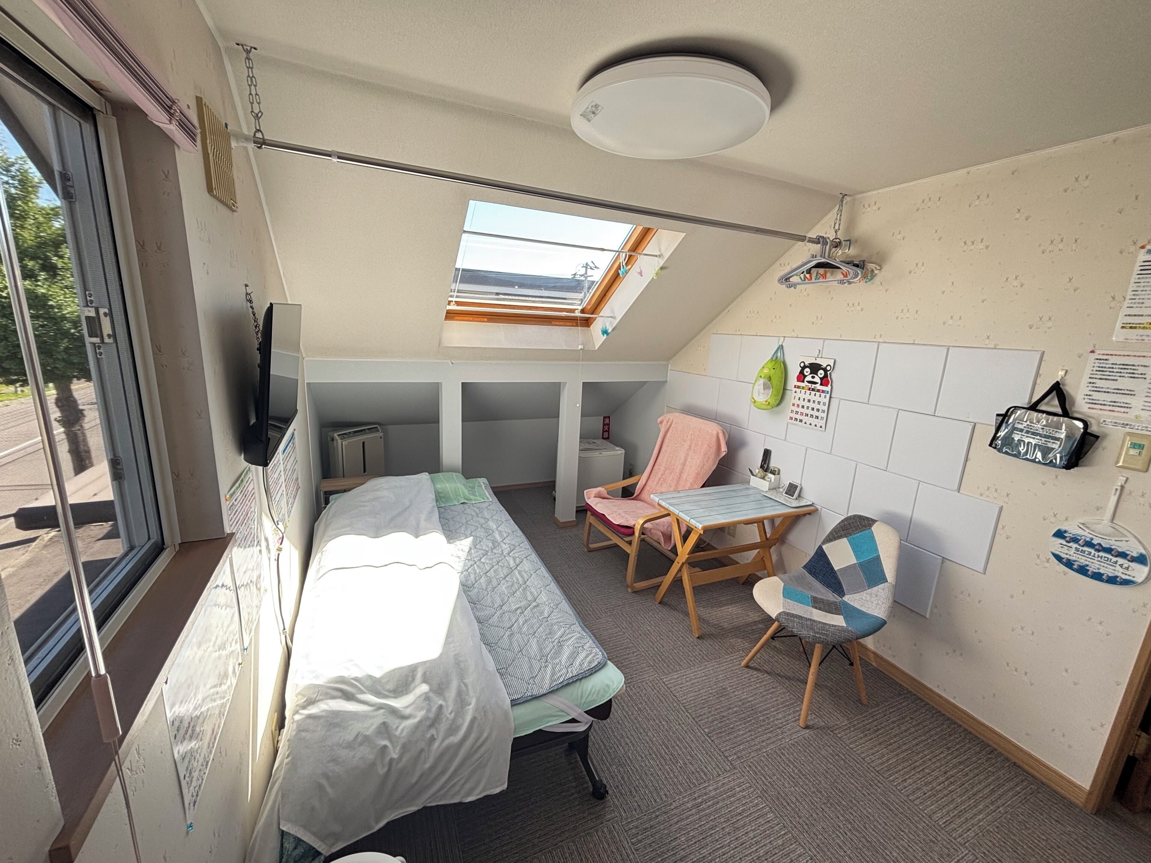 A compact room with a bed, a chair, a small table, a window, and a wall calendar.