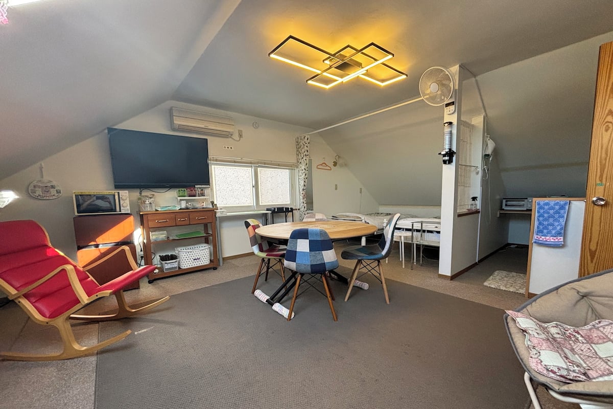 A compact, well-lit room with a dining table, chairs, a red rocking chair, a television, and a kitchenette area.
