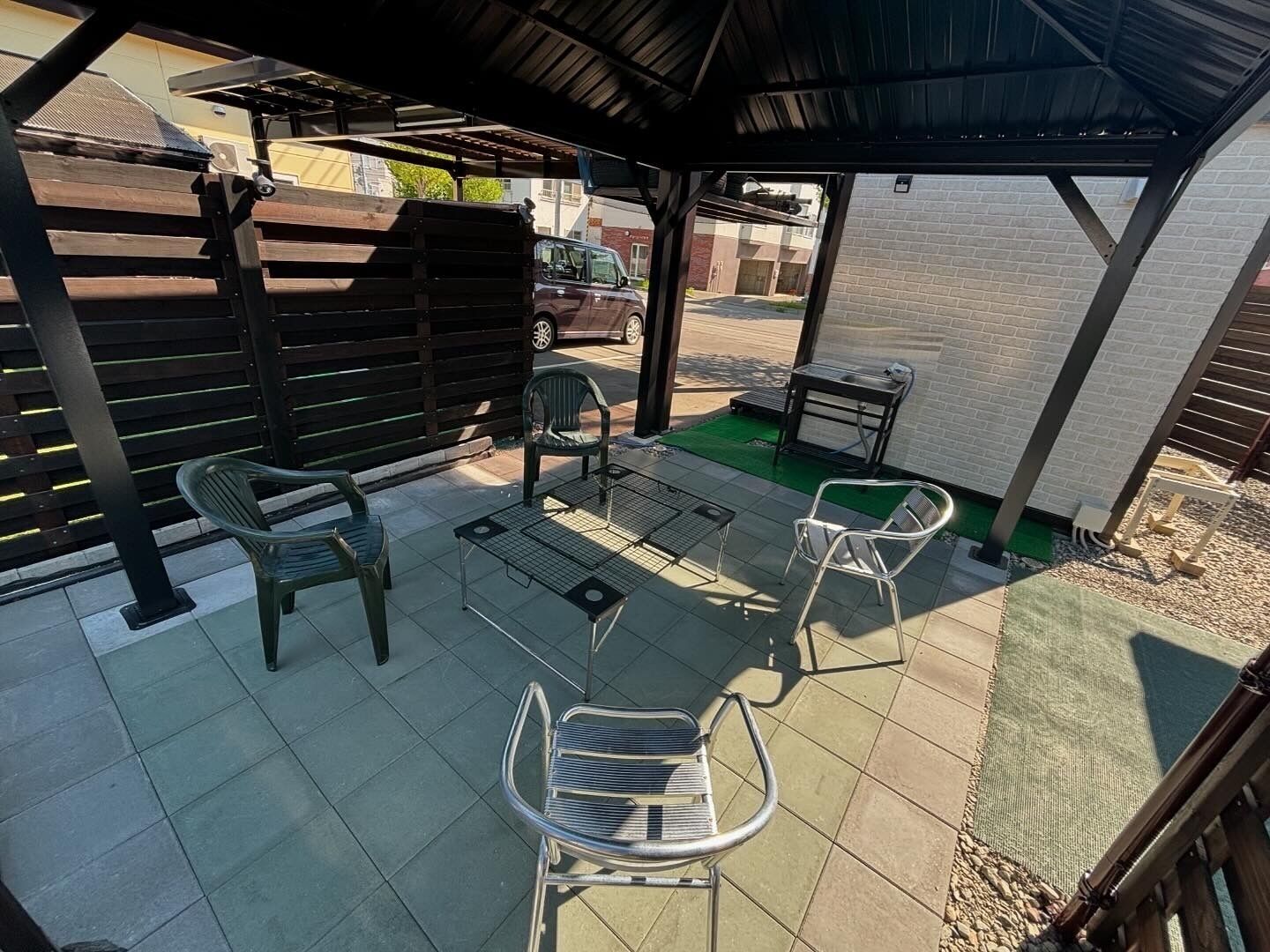 An outdoor patio area with a covered seating area, including metal chairs and a table, and a wooden fence in the background.