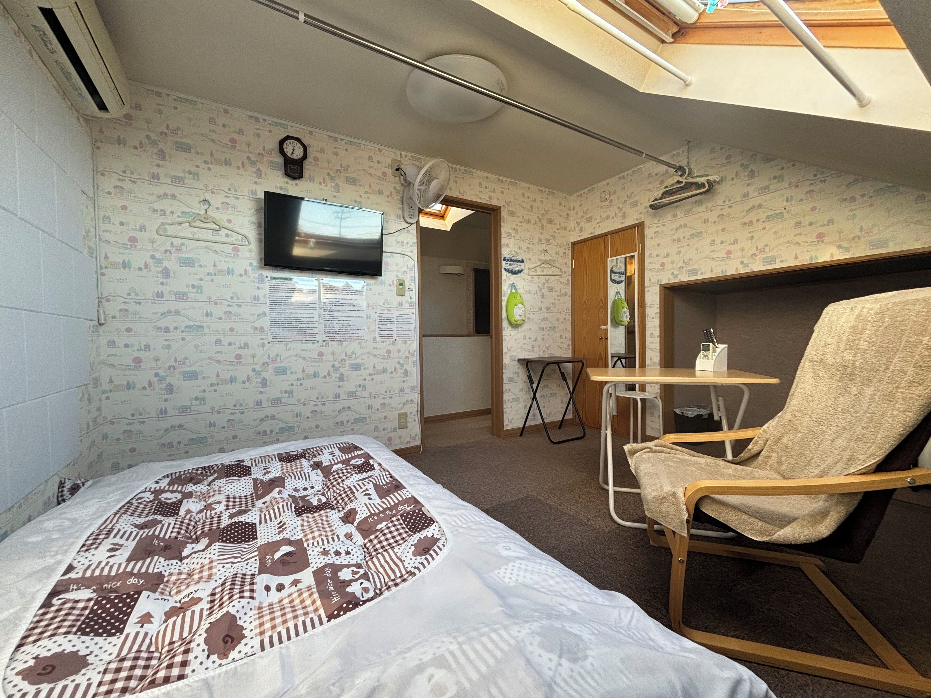 A bedroom with a bed, a chair, a small table, a TV, and a ceiling fan.