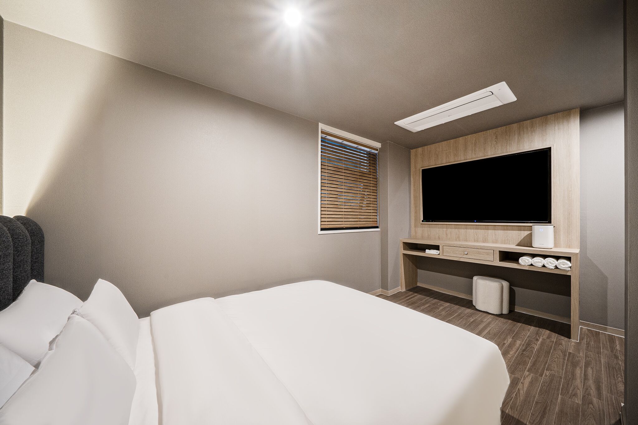 A hotel room with a large bed, a flat-screen TV mounted on the wall, a small desk with a chair, and a window with blinds.
