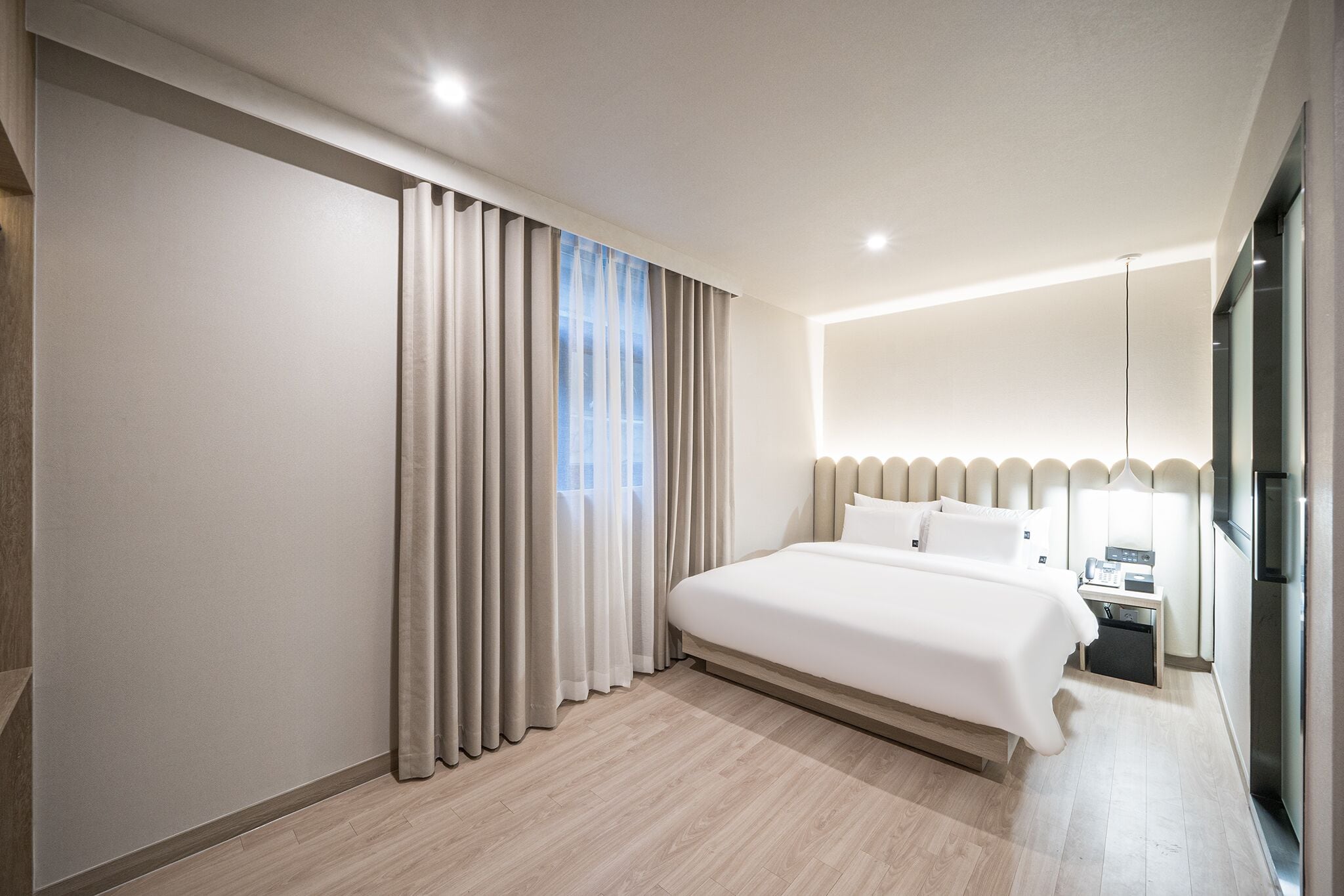 Quarto de hotel moderno com uma cama grande, mesa de cabeceira, escrivaninha embutida e janela com cortinas.