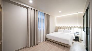 Quarto de hotel moderno com uma cama grande, mesa de cabeceira, escrivaninha embutida e janela com cortinas.